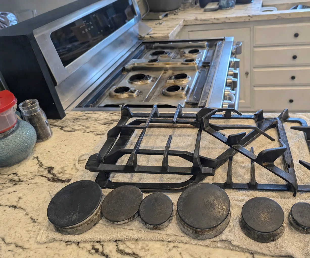 Burner caps of four different sizes lined up on granite next to their grates, the rangetop behind them with the burners bare